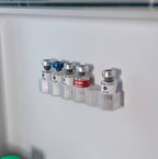 A set of five small vials with clear plastic holders mounted on a white fridge wall. The vials have silver caps, with three featuring blue, red, and black labels, and one with a white label.